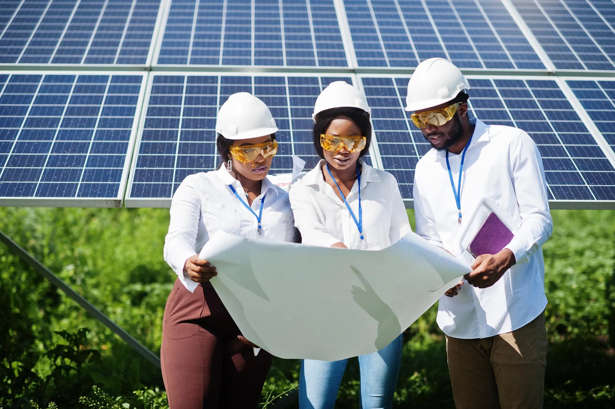 Grupo de ingenieros con cascos y gafas de seguridad revisando planos frente a paneles solares.