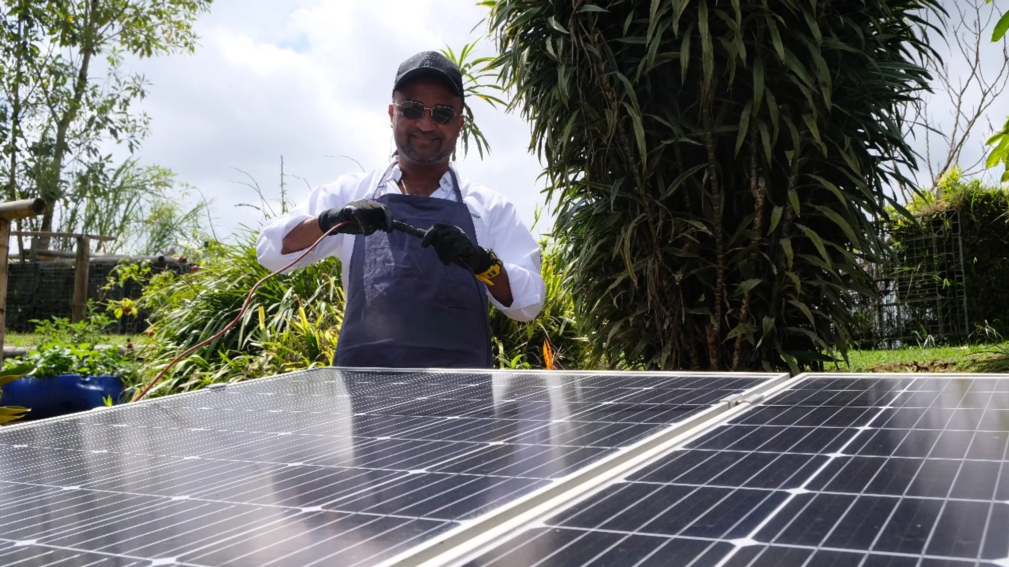 Hombre con guantes y gorra trabajando en paneles solares al aire libre con vegetación de fondo.